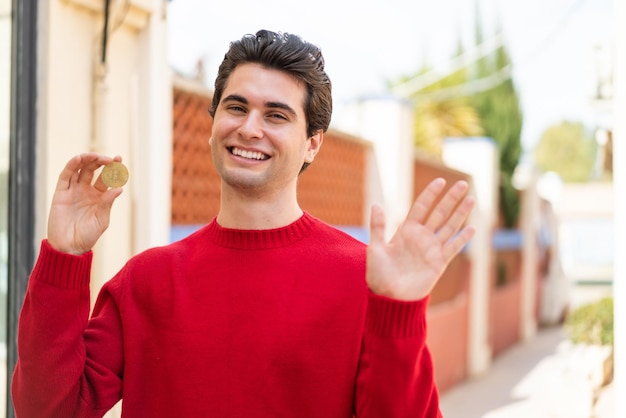 Young handsome man holding a Bitcoin at outdoors saluting with hand with happy expression