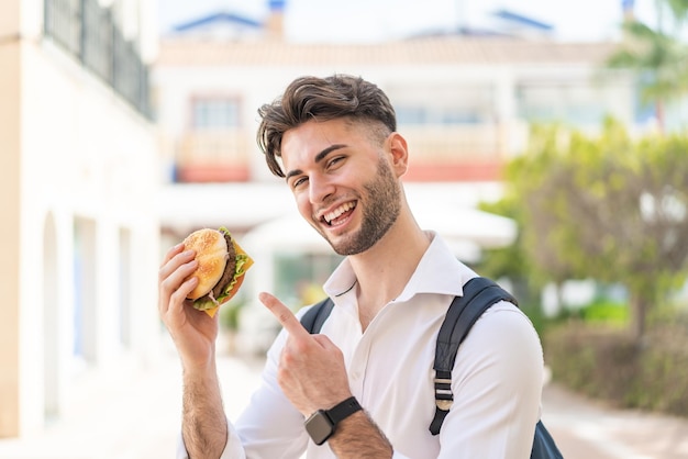 Молодой красивый мужчина держит гамбургер на улице и указывает на него
