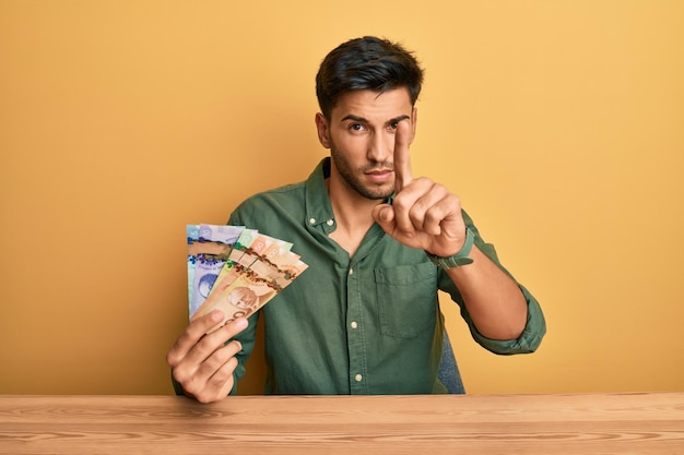 Young handsome man holding canadian dollars pointing with finger up and angry expression showing no gesture