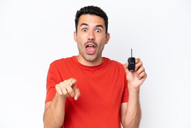 Young handsome man holding car keys over isolated white background surprised and pointing front