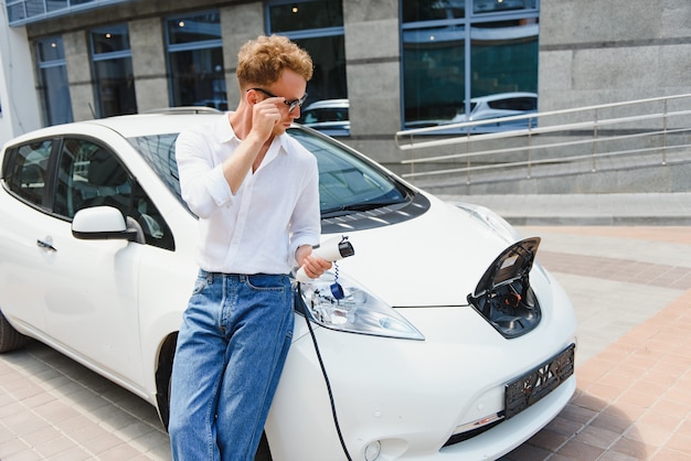 彼の新しい車の近くに立って、満足しているように見える充電ステーションポイントで充電ケーブルを保持している若いハンサムな男。