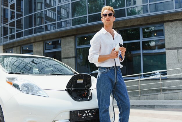 Young handsome man holding charging cable at electric charging station point standing near his new car , looking satisfied.