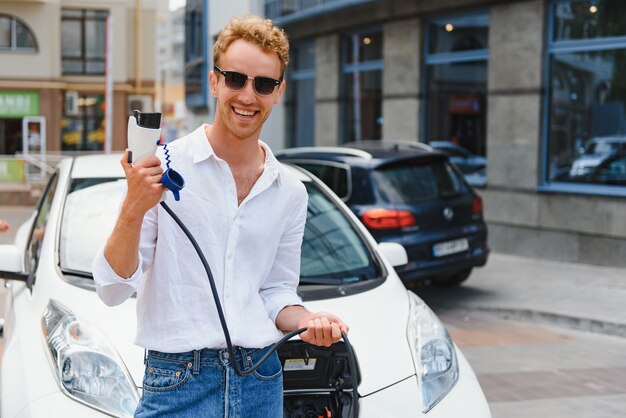 彼の新しい車の近くに立って、満足しているように見える充電ステーションポイントで充電ケーブルを保持している若いハンサムな男。