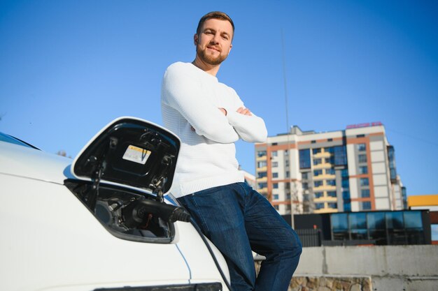 Young handsome man holding charging cable at electric charging station point standing near his new car.