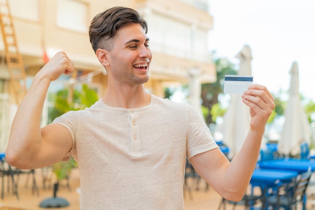 Young handsome man holding a credit card at outdoors celebrating a victory