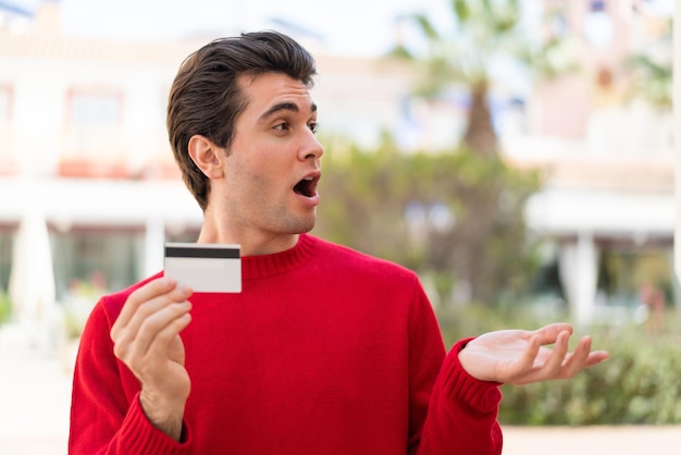 Young handsome man holding a credit card at outdoors with surprise facial expression