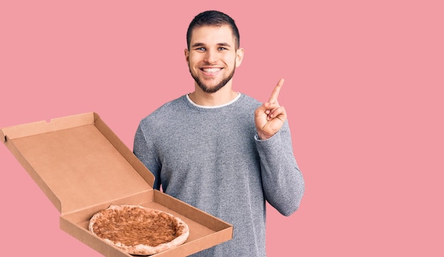 Young handsome man holding delivery italian pizza surprised with an idea or question pointing finger with happy face number one