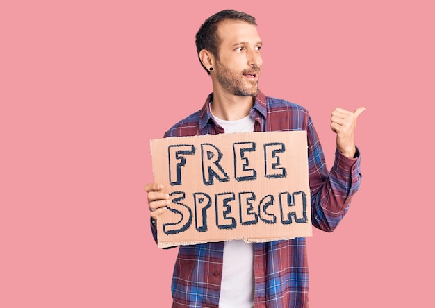 Young handsome man holding free speech banner pointing thumb up to the side smiling happy with open mouth