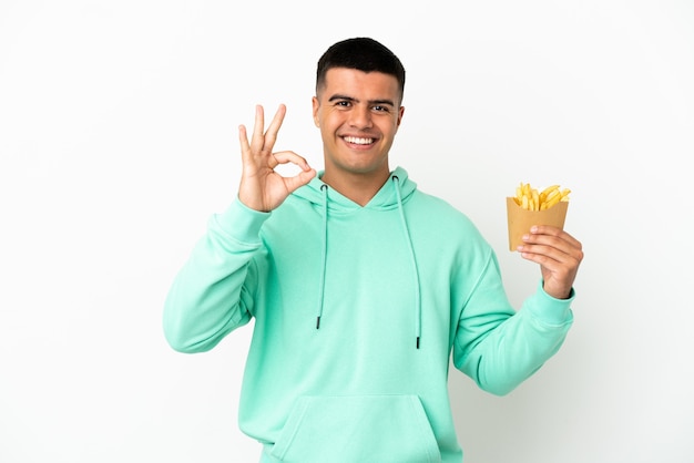 Young handsome man holding fried chips over isolated white background showing ok sign with fingers