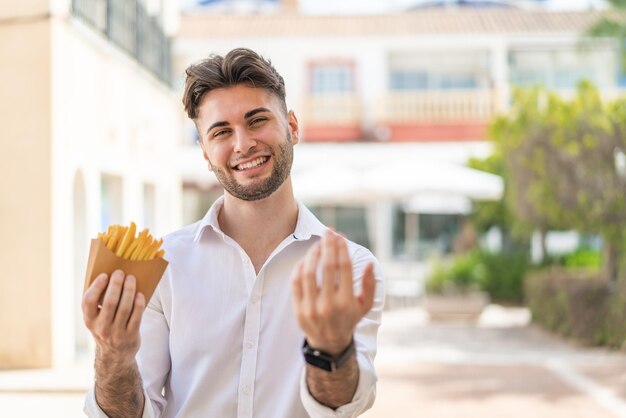 Foto giovane bell'uomo che tiene patatine fritte all'aperto invitando a venire con la mano felice che tu sia venuto