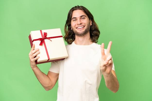 Young handsome man holding a gift over isolated background smiling and showing victory sign