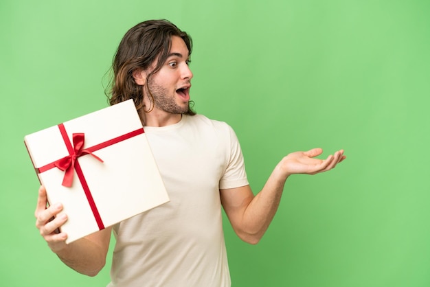 Young handsome man holding a gift over isolated background with surprise facial expression