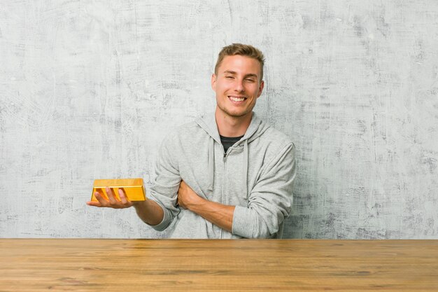 Young handsome man holding a gold ingot on a table laughing and having fun.