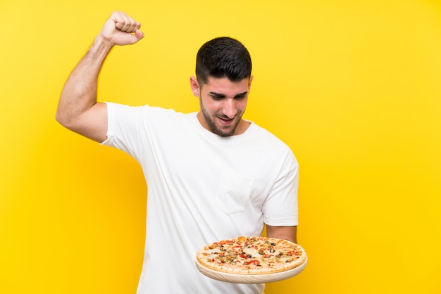 Young handsome man holding a pizza over isolated yellow wall celebrating a victory