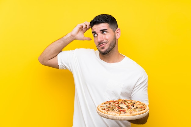 Young handsome man holding a pizza over isolated yellow wall having doubts and with confuse face expression