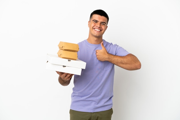 Молодой красавец держит пиццу и гамбургеры на изолированном белом фоне, показывая большой палец вверх