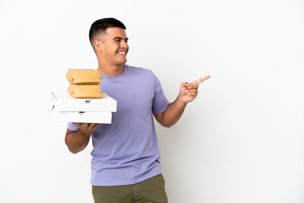 Young handsome man holding pizzas and burgers over isolated white background pointing finger to the side and presenting a product