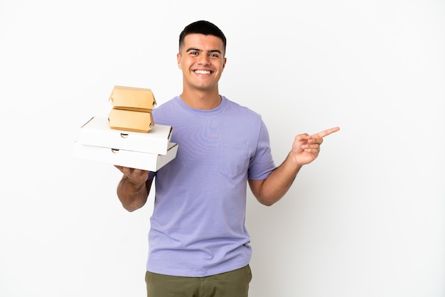 Молодой красавец держит пиццу и гамбургеры на изолированном белом фоне, указывая пальцем в сторону