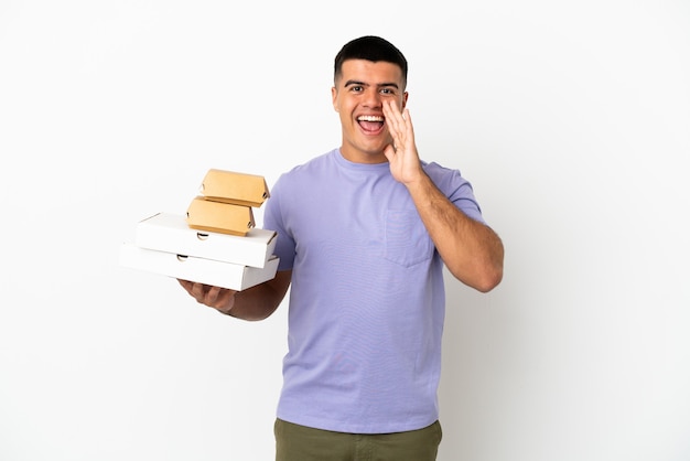 Young handsome man holding pizzas and burgers over isolated white background shouting with mouth wide open