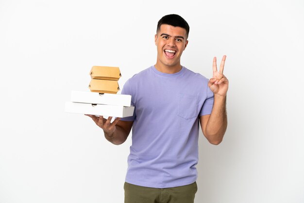 Young handsome man holding pizzas and burgers over isolated white background smiling and showing victory sign