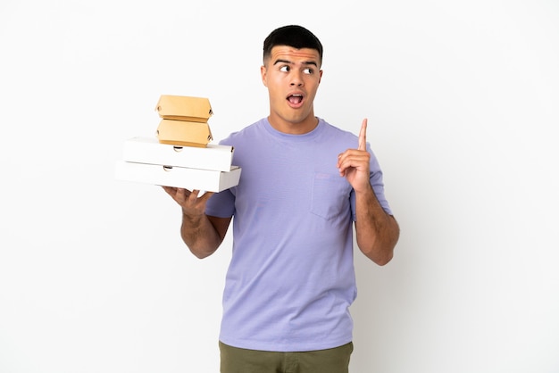 Photo young handsome man holding pizzas and burgers over isolated white background thinking an idea pointing the finger up