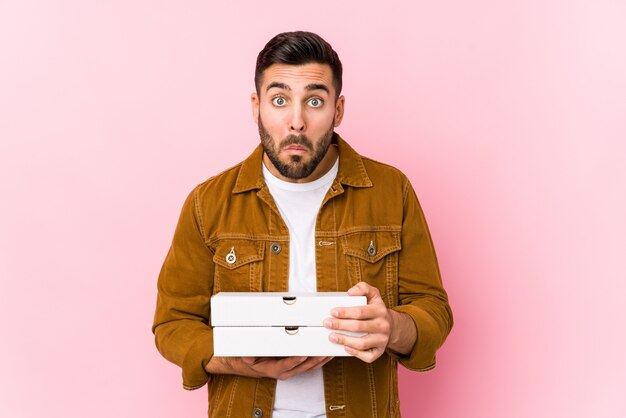 Young handsome man holding pizzas shrugs shoulders and open eyes confused.