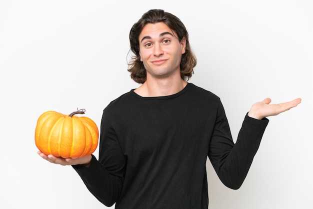 Giovane uomo bello che tiene una zucca isolata su sfondo bianco che ha dubbi mentre alza le mani