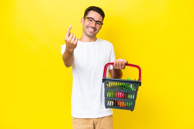 Young handsome man holding a shopping basket full of food over isolated yellow background doing coming gesture