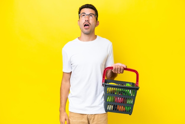 Young handsome man holding a shopping basket full of food over isolated yellow background looking up and with surprised expression