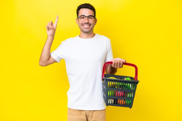 Young handsome man holding a shopping basket full of food over isolated yellow background pointing up a great idea