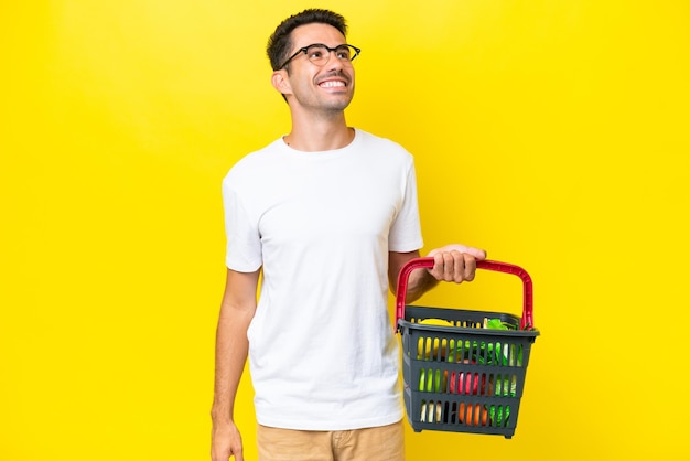 Young handsome man holding a shopping basket full of food over isolated yellow background thinking an idea while looking up