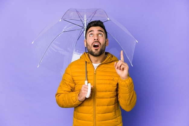 Young handsome man holding an umbrella isolated pointing upside with opened mouth.