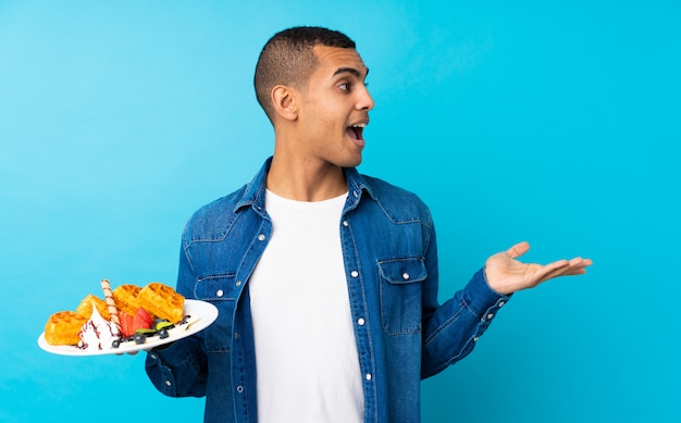 Young handsome man holding waffles over isolated blue wall with surprise facial expression