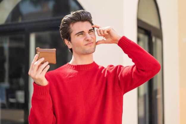 Young handsome man holding a wallet at outdoors having doubts and with confuse face expression