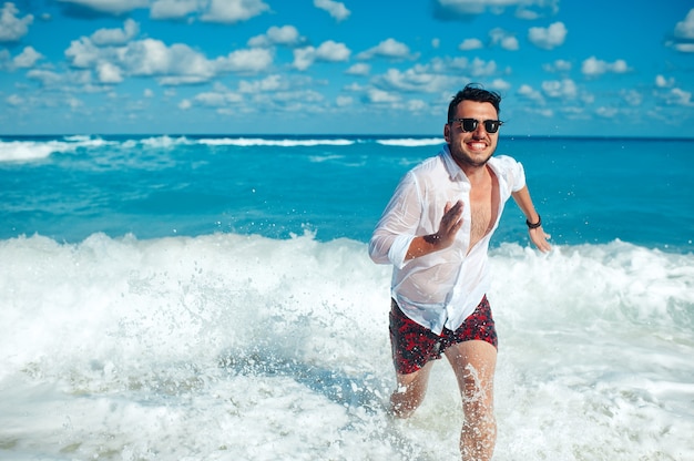 Фото Молодой красавец в белой рубашке и солнцезащитных очках бежит по морским волнам