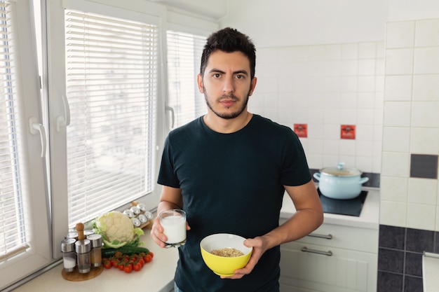 Young handsome man is having breakfast in the kitchen at home. The guy prepared himself oatmeal and a glass of milk for breakfast.