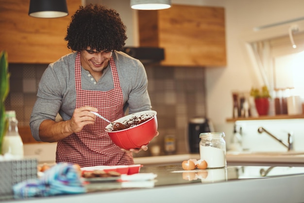 写真 若いハンサムな男は彼の友人のために彼の台所でクッキーを作っています。彼はチョコレート生地を型に流し込んでいます。