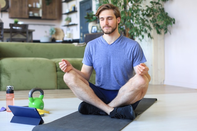 Young handsome man is practicing yoga in lotus pose sitting on the floor at home during quarantine. Sport is foundation of healthy life.