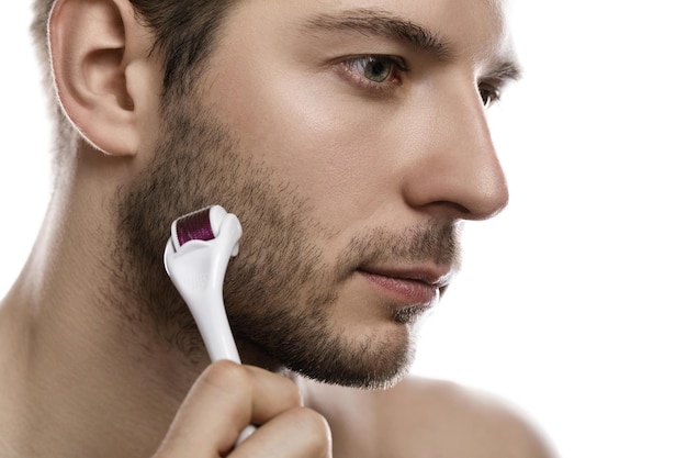 Young handsome man is using derma roller for beard growth on white background