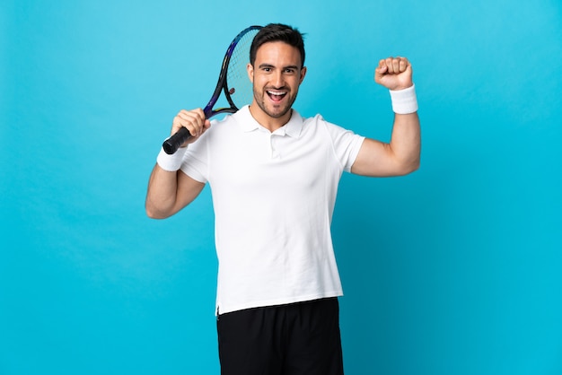 Young handsome man isolated on blue background playing tennis and celebrating a victory