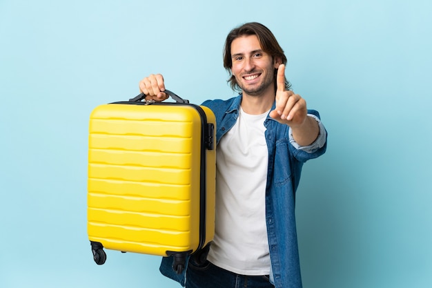 Young handsome man isolated on blue background in vacation with travel suitcase and counting one