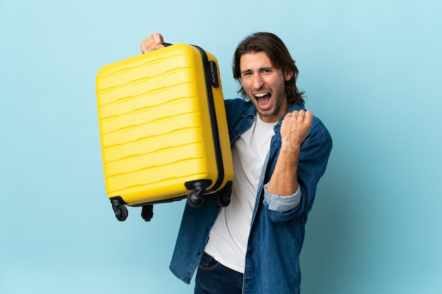 Young handsome man isolated on blue background in vacation with travel suitcase