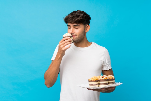 Young handsome man over isolated blue holding mini cakes