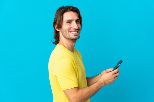 Young handsome man isolated on blue sending a message or email with the mobile