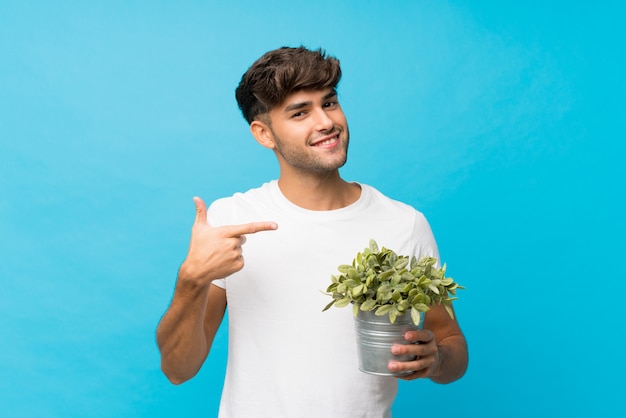 Young handsome man over isolated blue  taking a flowerpot and pointing it