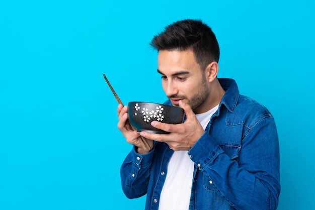 Young handsome man over isolated blue wall holding a bowl of noodles with chopsticks and eating it