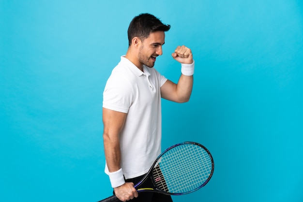 Young handsome man isolated on blue wall playing tennis and celebrating a victory