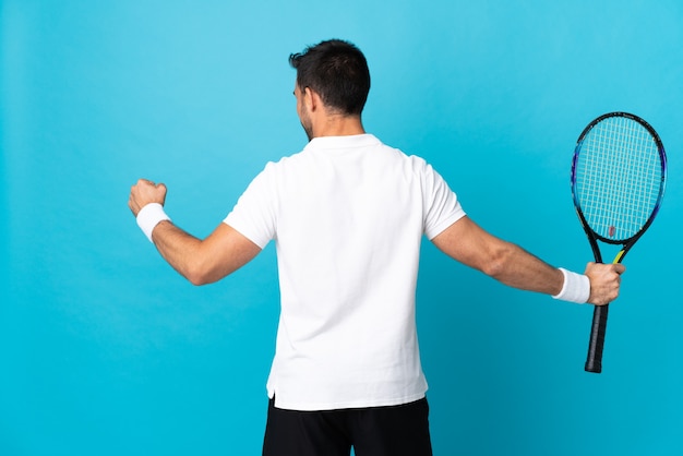 Young handsome man isolated on blue wall playing tennis and celebrating a victory