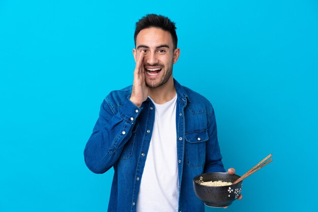 Young handsome man over isolated blue wall shouting with mouth wide open while holding a bowl of noodles with chopsticks