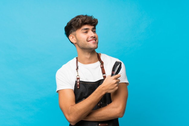 Young handsome man over isolated blue with hairdresser or barber dress and holding hair cutting machine